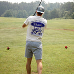 Person on a golf course wearing a white t-shirt with branding and gray shorts, holding a golf club.